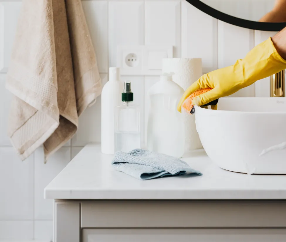 A hand wearing a yellow rubber glove cleans a white bowl on a bathroom counter cluttered with bottles and a towel.
