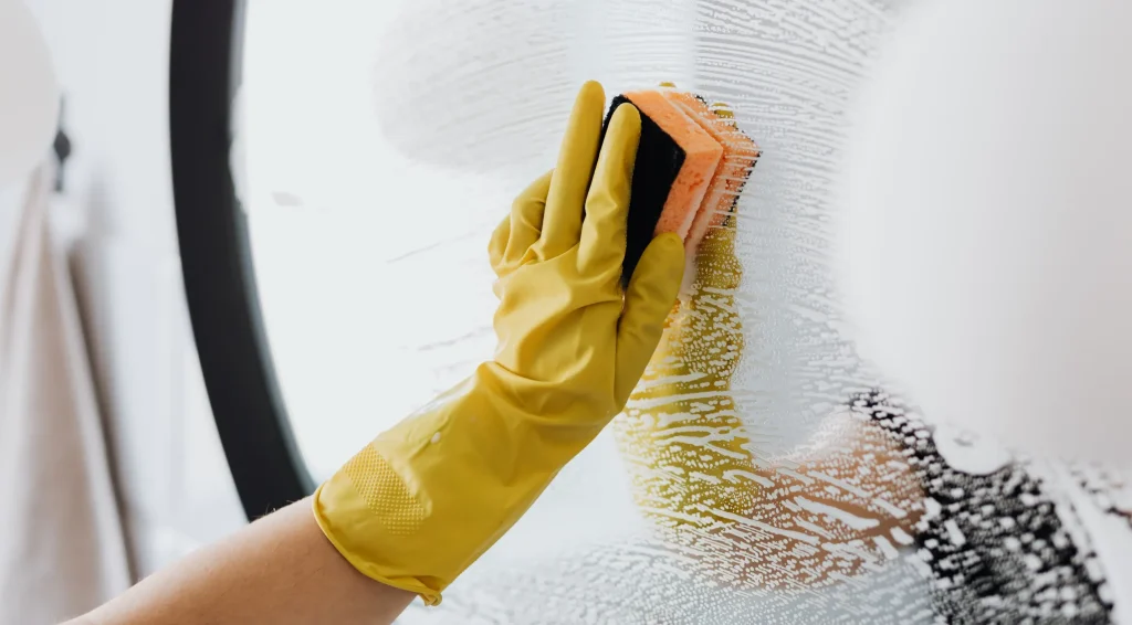 A hand wearing a yellow glove scrubs a glass surface with a sponge, creating foam and streaks while cleaning.