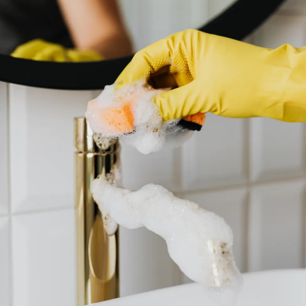A person in a yellow glove scrubs a gold faucet with a soapy sponge, cleaning a bathroom sink area.