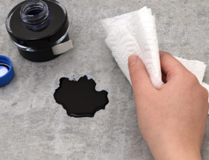 Hand using a paper towel to clean up spilled black ink from a bottle on a gray surface