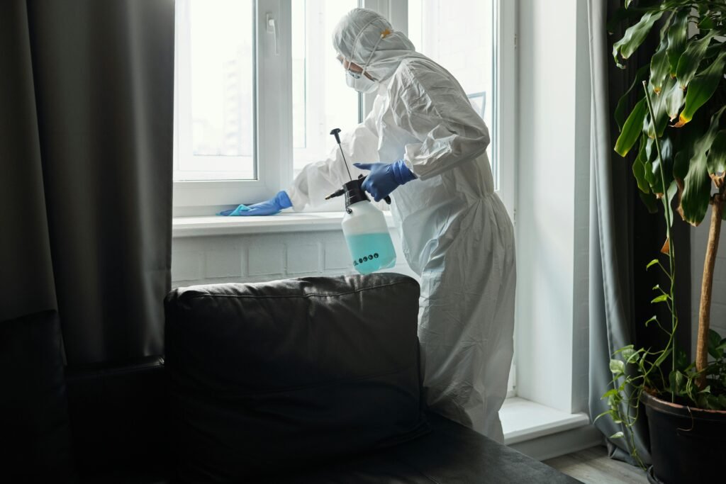 A person in a full protective suit sprays disinfectant while cleaning a window in a modern living room.