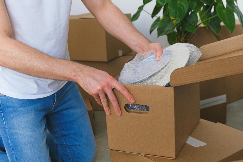 A person unpacks a fragile item covered in bubble wrap from a cardboard box, surrounded by more boxes and a potted plant.
