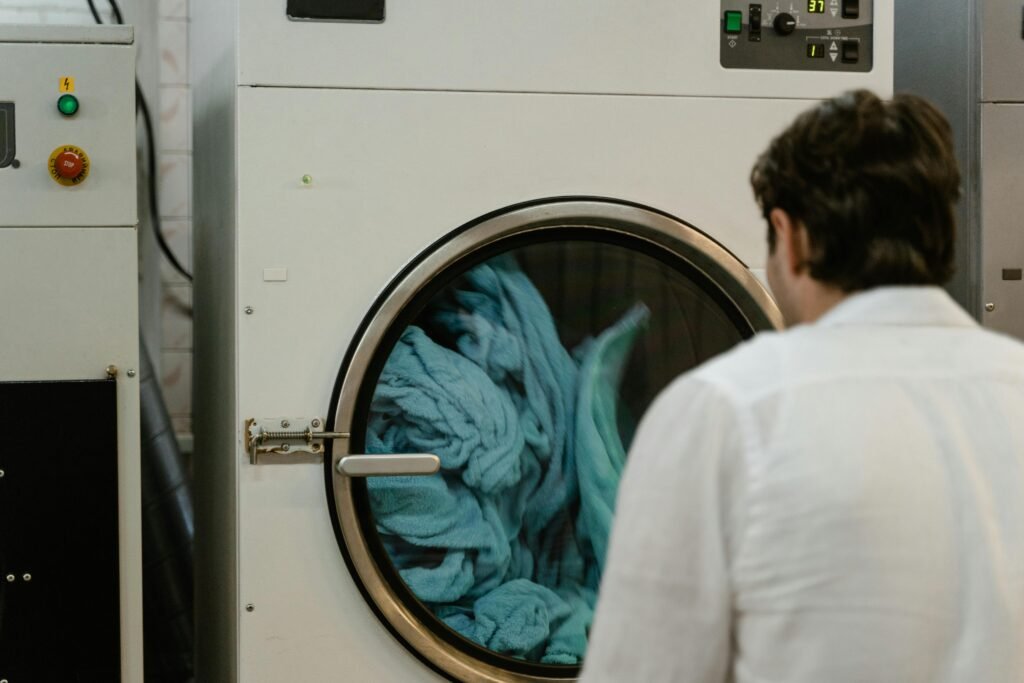 A man observes a large laundry machine as it churns blue towels inside a commercial laundry facility.
