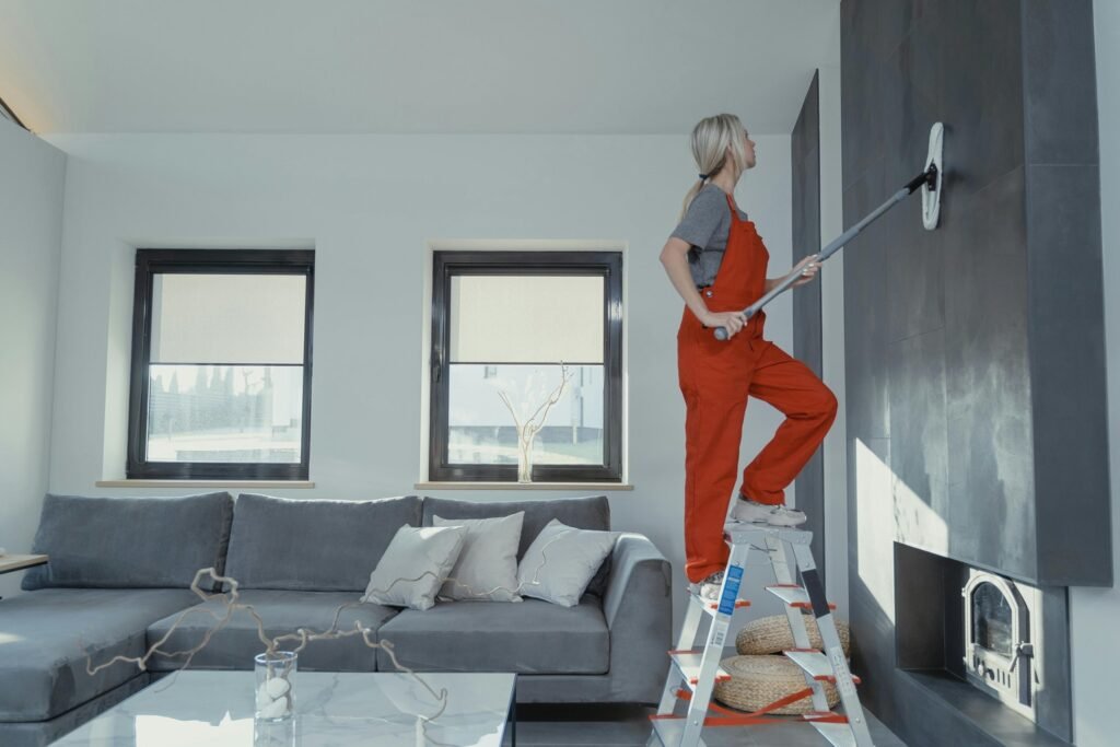 A person in red overalls paints a dark wall while standing on a ladder in a modern living room with gray furniture.