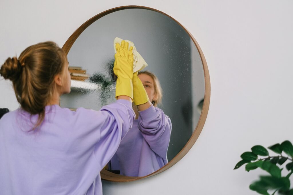 A person wearing yellow cleaning gloves wipes a round mirror with a cloth, surrounded by indoor plants and shelves.