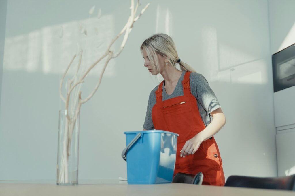 A woman in an orange overall prepares to clean a modern room, featuring a vase with dried twigs on the table.