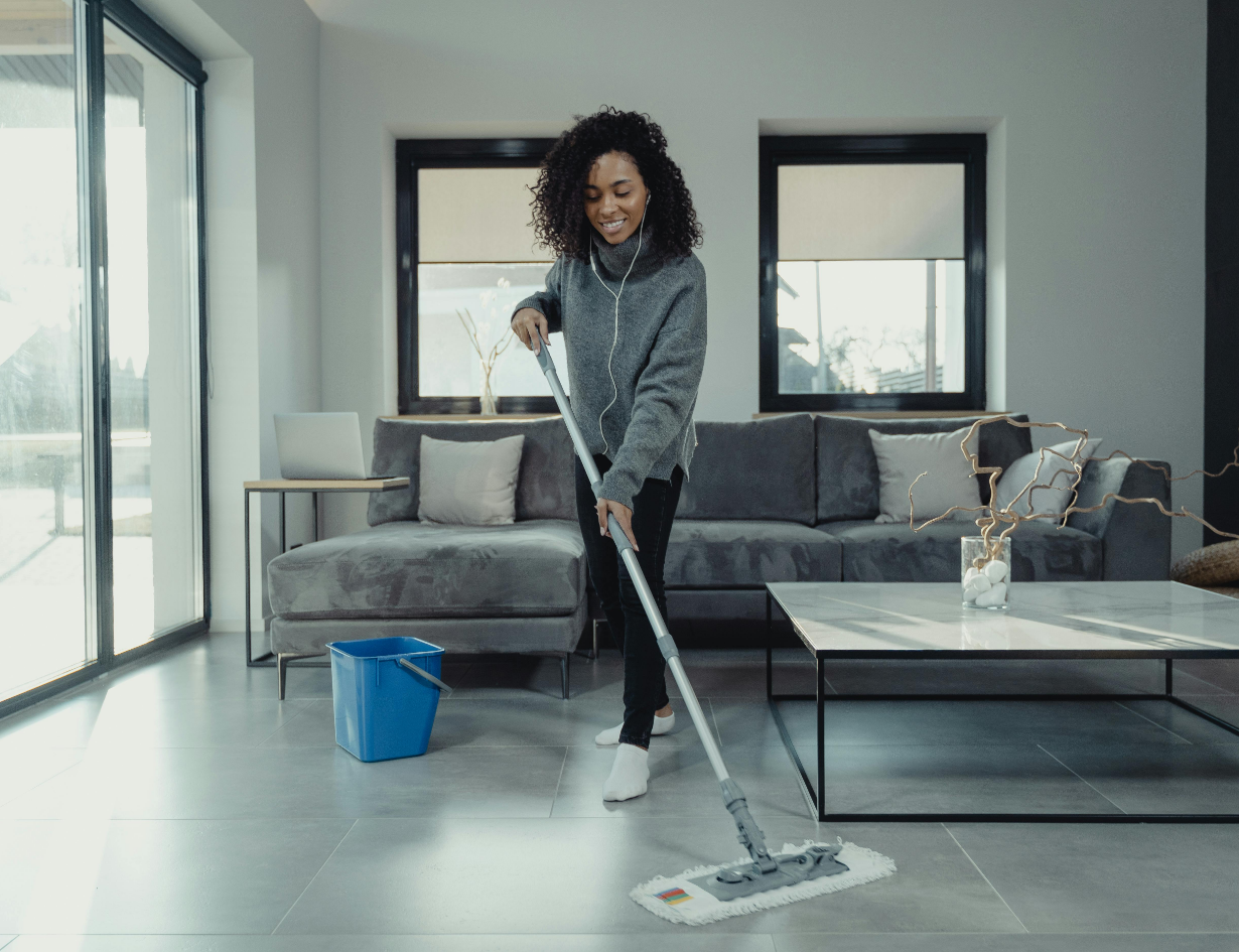A person mops the floor in a modern living room with large windows, a gray sofa, and a blue bucket nearby.