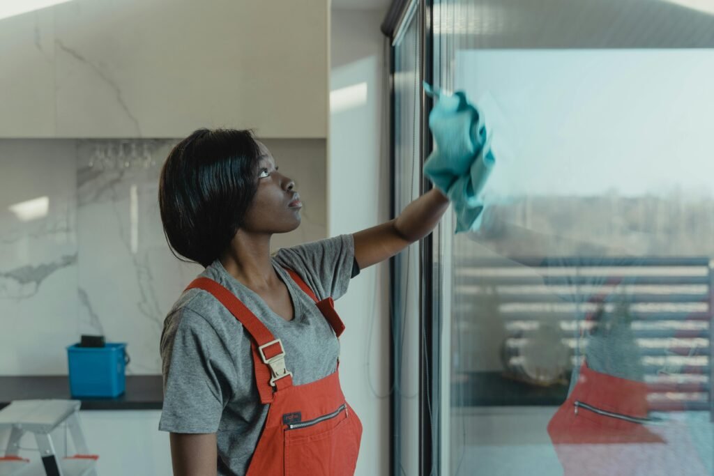 A person in red overalls wipes a large glass window with a blue cloth, with a modern kitchen in the background.
