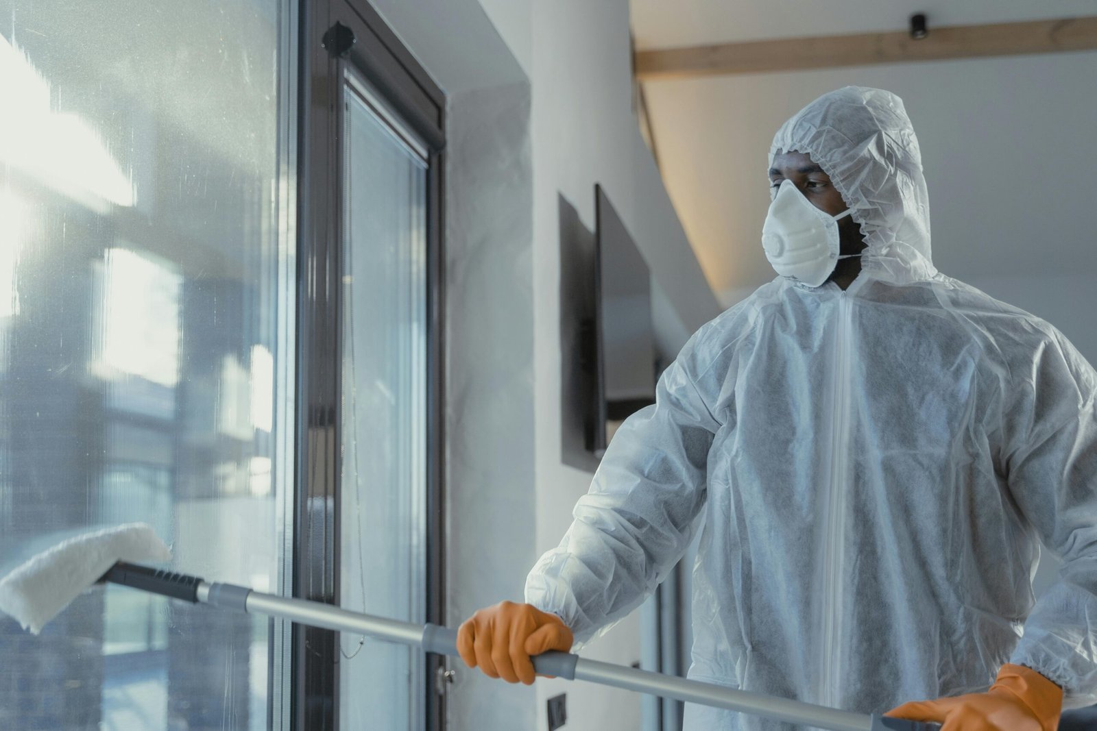 A person in a protective suit and orange gloves cleans a large window with a mop in a bright, modern indoor space.