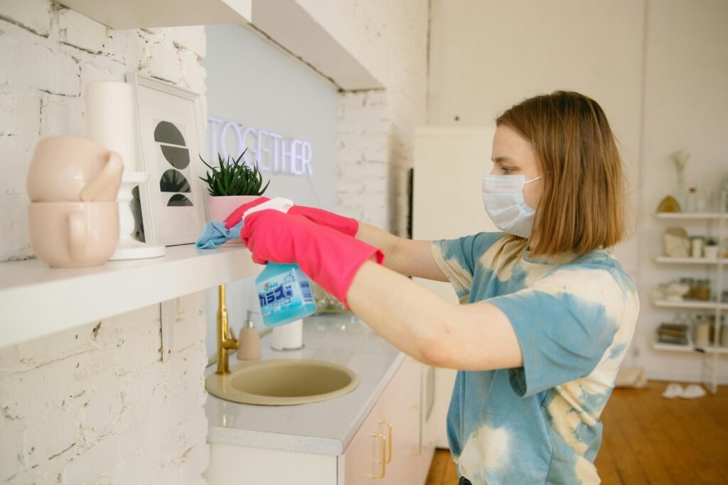 A person in a tie-dye shirt cleans a white shelf adorned with plants, cups, and artwork, using a spray cleaner and cloth.