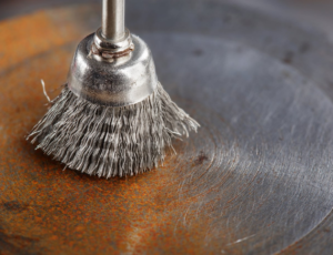 A close-up of a metal brush cleaning a rusted surface, revealing shiny metal beneath the debris and oxidation.