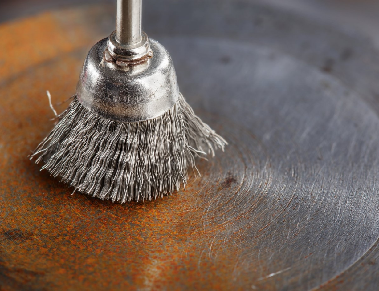 A close-up of a metal brush cleaning a rusted surface, revealing shiny metal beneath the debris and oxidation.