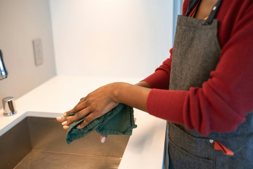 A person in an apron wipes their hands with a green cloth near a stainless steel sink in a modern kitchen.