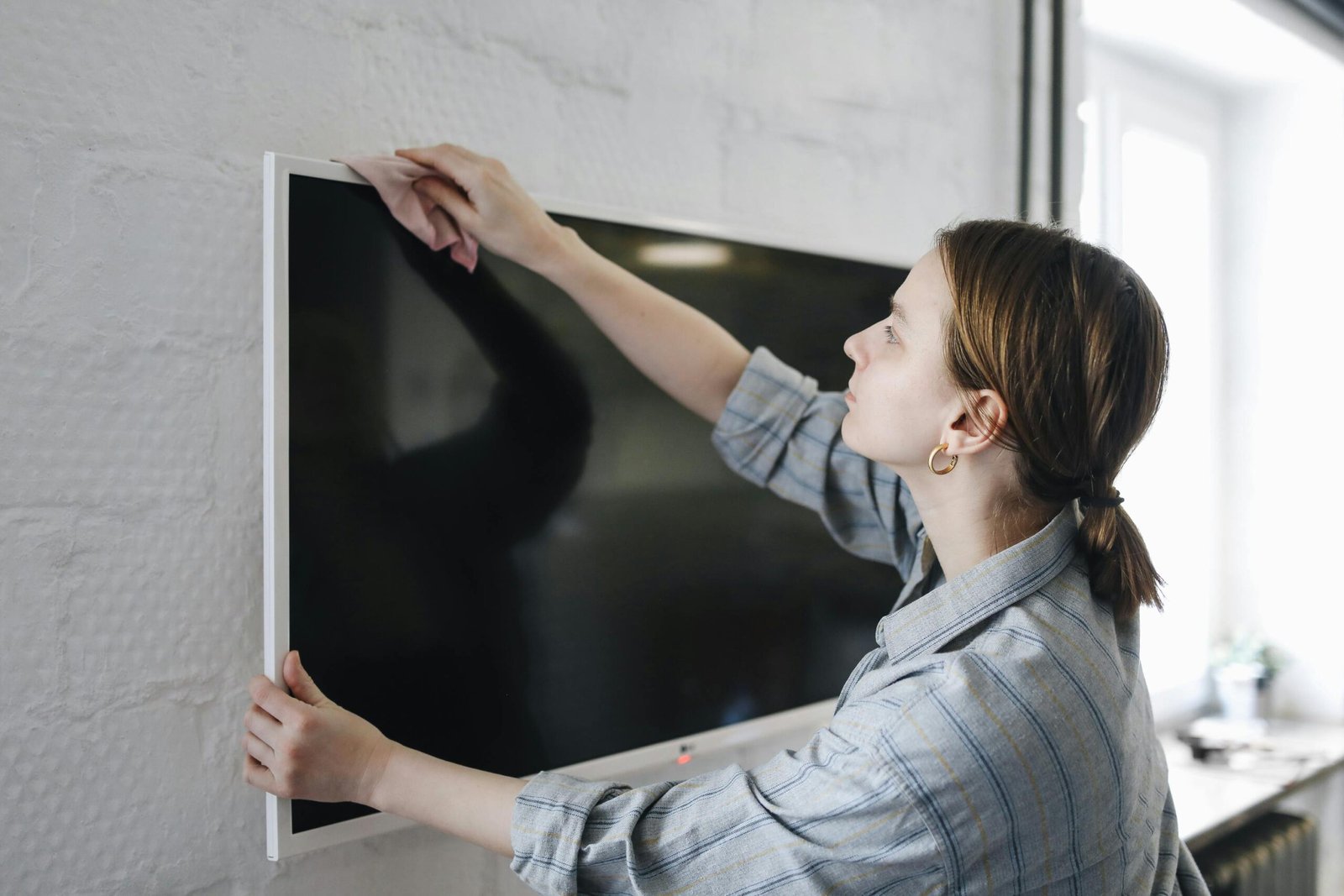 A person wearing a plaid shirt is cleaning a large flat-screen television mounted on a white brick wall.