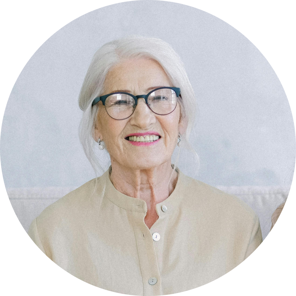 An elderly woman with long, silver hair wears a light beige button-up top, seated against a soft, muted background.
