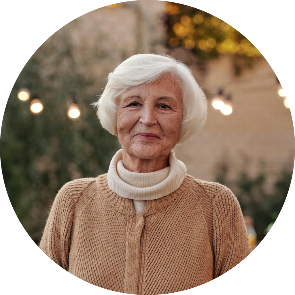 An elderly woman with short white hair wears a beige cardigan and a white turtleneck, standing in a softly lit outdoor setting.