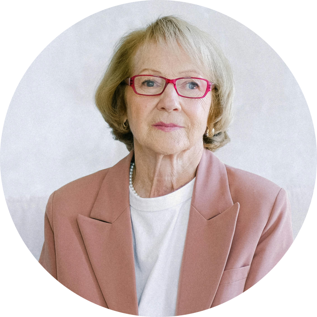 An elderly woman in a pink blazer and white shirt poses against a soft, light background, exuding elegance and calmness.