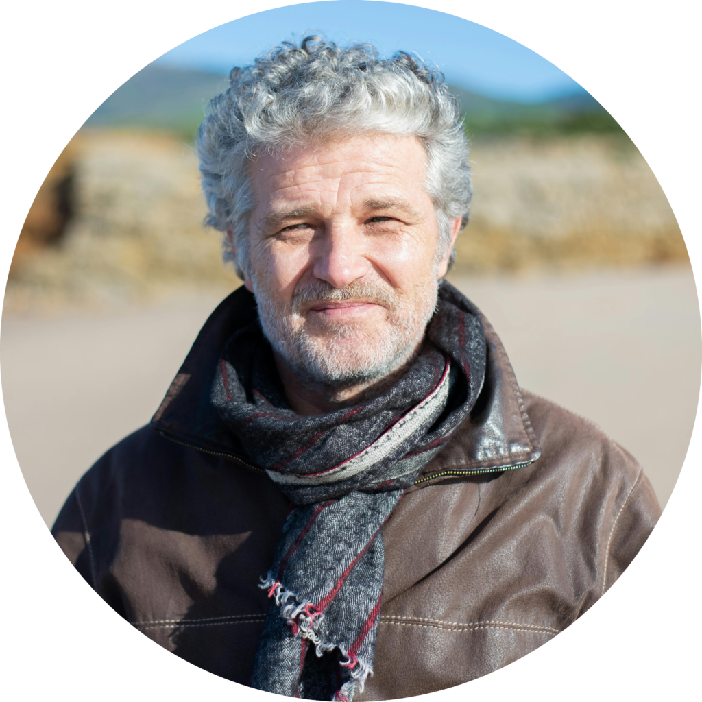 A man with curly gray hair wears a leather jacket and a patterned scarf, standing outdoors with a blurred background.