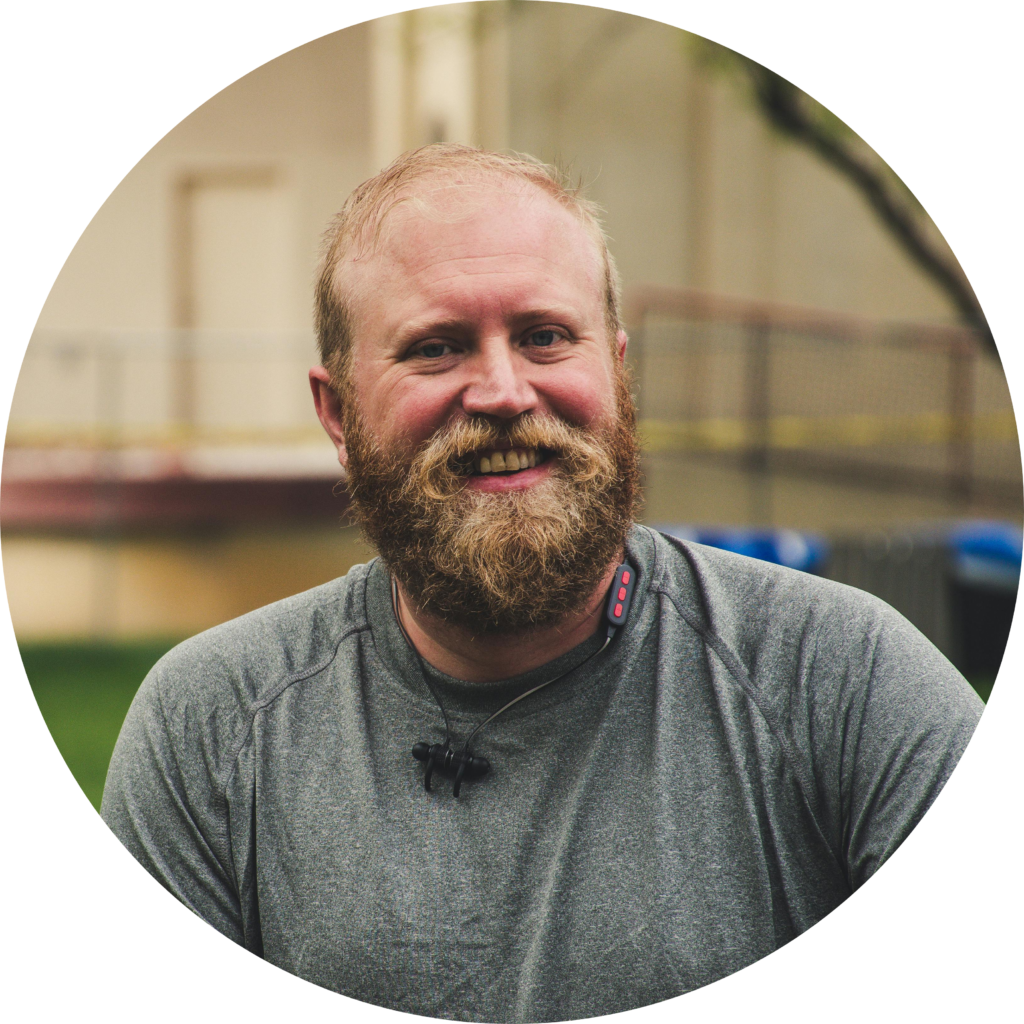 Smiling bearded man in grey athletic shirt with headset, outdoors near building with blurred background